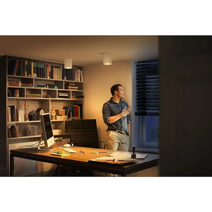 Philips Hue LED-Deckenstrahler White Ambiance Pillar Erweiterung - L x B x H: 10,3 x 10,3 x 12 cm, Weiß, Neutralweiß - Mann steht am Fenster in einem Raum mit zwei zylindrischen Deckenleuchten, die ein warmes Licht spenden.