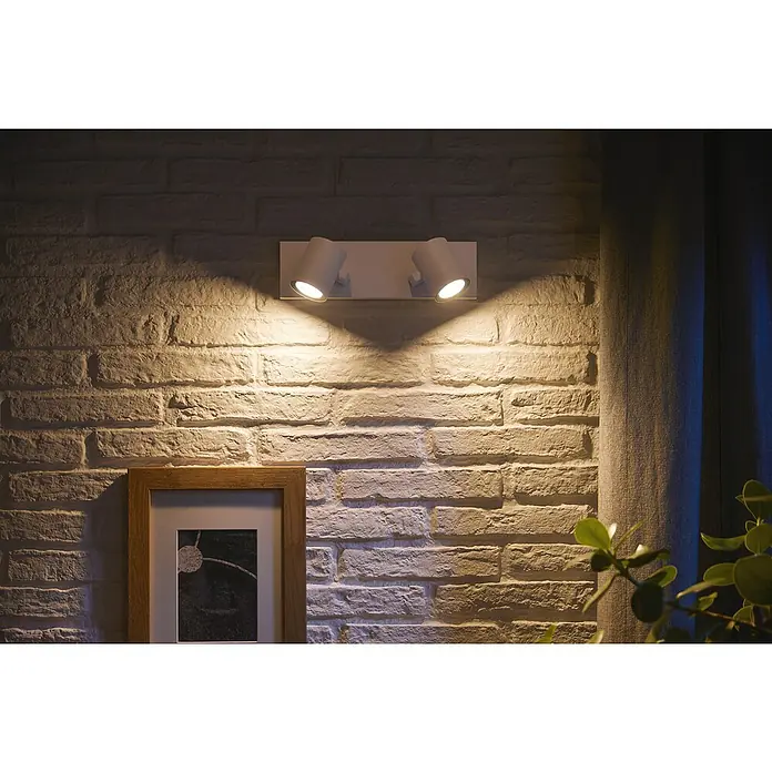 Philips Hue LED-Deckenstrahler White and Color Ambience Argenta - 4,2 W, L x B x H: 30,5 x 8,9 x 12,6 cm, Weiß, RGBW - Wandleuchte mit zwei Strahlern, die ein warmes Licht auf eine Ziegelwand wirft und eine gemütliche Atmosphäre schafft.