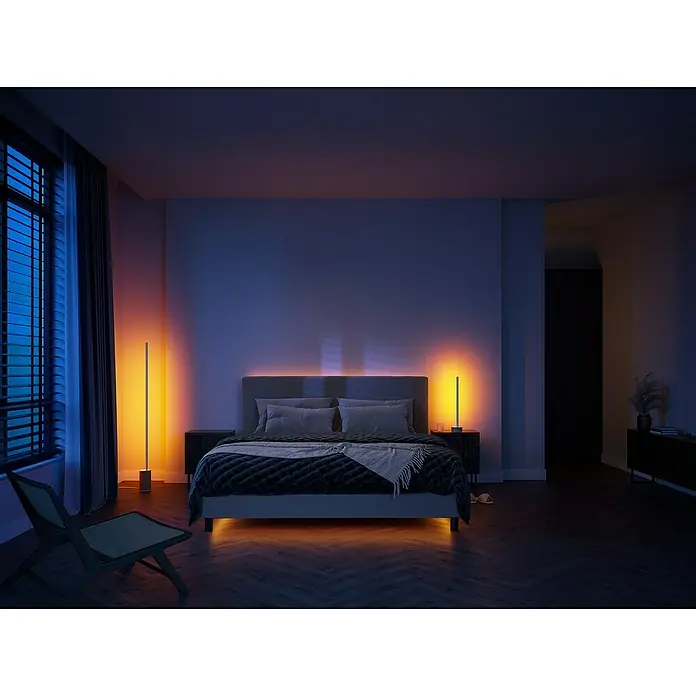 Philips Hue LED-Tischleuchte Signe Oak - 12 W, L x B x H: 11,15 x 11,15 x 55,3 cm, Weiß, RGBW - Stimmungsaufnahme eines Schlafzimmers mit einem beleuchteten Bett und zwei leuchtenden Lampen, die eine warme Atmosphäre erzeugen.