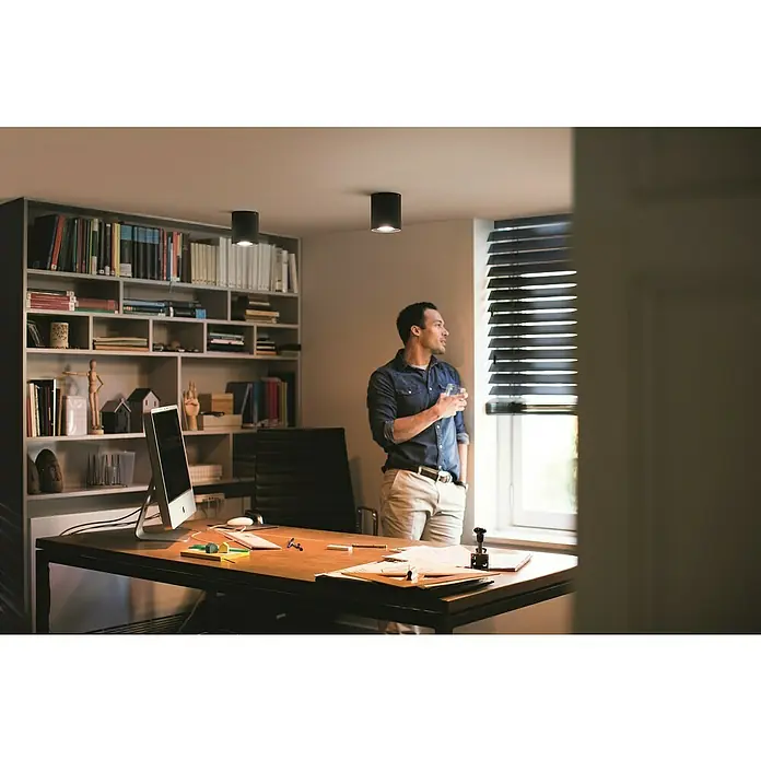 Philips Hue LED-Deckenstrahler White Ambiance Pillar Erweiterung - L x B x H: 10,3 x 10,3 x 12 cm, Schwarz, Neutralweiß - Ein Mann steht mit einem Glas in einem Büro mit schwarzen Deckenleuchten und einem Bücherregal. Stilvolle Beleuchtung für modernes Arbeiten.