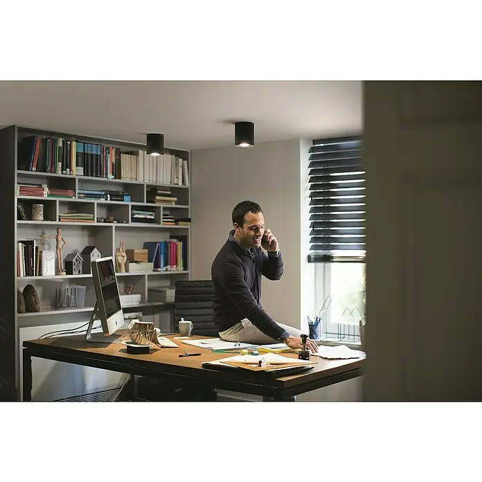 Philips Hue LED-Deckenstrahler White Ambiance Pillar Erweiterung - L x B x H: 10,3 x 10,3 x 12 cm, Schwarz, Neutralweiß - Ein Mann sitzt auf einem Schreibtisch und telefoniert mit einem Handy in einem modern eingerichteten Arbeitszimmer mit Deckenleuchten.