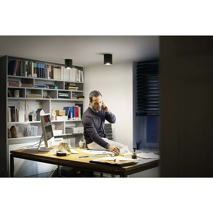 Philips Hue LED-Deckenstrahler White Ambiance Pillar - 4,2 W, L x B x H: 10,3 x 10,3 x 12 cm, Schwarz, Neutralweiß - Ein Mann sitzt auf einem Schreibtisch unter zylindrischen Deckenleuchten und telefoniert. Die Leuchten sorgen für eine angenehme Atmosphäre im Raum.