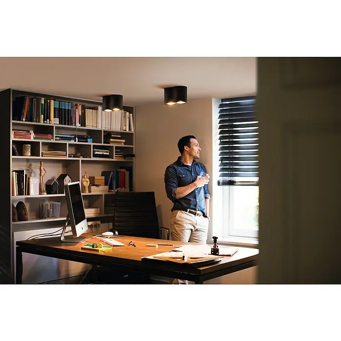 Philips Hue LED-Deckenstrahler White Ambience Pillar - 4,2 W, L x B x H: 19,3 x 10,3 x 12 cm, Weiß, Neutralweiß - Ein Mann steht am Fenster in einem Büro mit einer modernen Deckenleuchte, die für eine angenehme Beleuchtung sorgt.