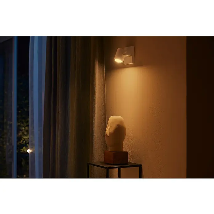 Philips Hue LED-Deckenstrahler White and Color Ambience Argenta - 4,2 W, L x B x H: 9 x 10,63 x 19 cm, Weiß, RGBW, Anzahl Leuchtmittel: 1 Stk. - Eine Nahaufnahme einer Wandleuchte, die eine sanfte, warme Beleuchtung in einem gemütlichen Raum erzeugt und eine Skulptur hervorhebt.