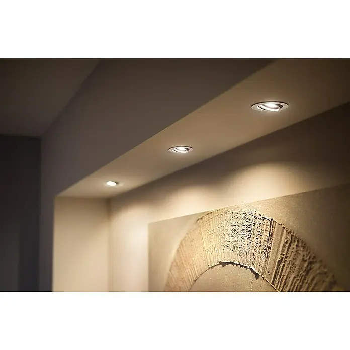 Philips Hue LED-Einbauleuchten-Set rund White Ambience Recessed Milliskin - 4,2 W, Ø x H: 9 x 10 cm, Alu, 3 Stk., Kaltweiß - Deckenleuchten in einem Raum mit Kunstwerk an der Wand. Die Leuchten sind kreisförmig und geben ein warmes, gerichtetes Licht ab.