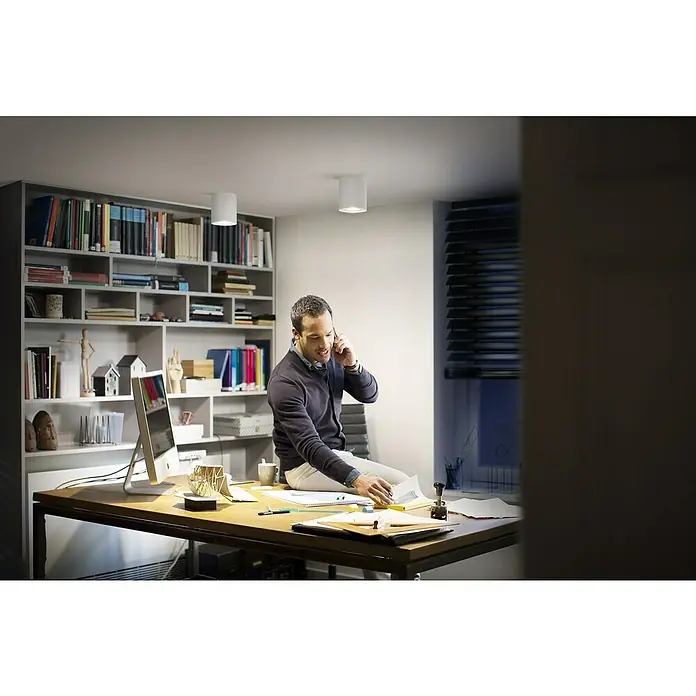 Philips Hue LED-Deckenstrahler White Ambiance Pillar Erweiterung - L x B x H: 10,3 x 10,3 x 12 cm, Weiß, Neutralweiß - Der Mann sitzt auf dem Schreibtisch, telefoniert und schaut sich seine Unterlagen unter einer Deckenleuchte an.