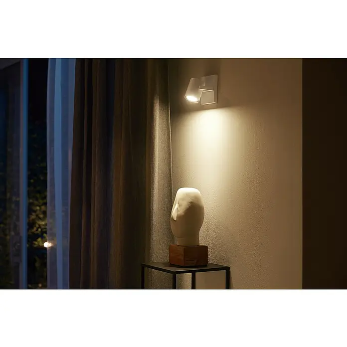 Philips Hue LED-Deckenstrahler White and Color Ambience Argenta - 4,2 W, L x B x H: 9 x 10,63 x 19 cm, Weiß, RGBW, Anzahl Leuchtmittel: 1 Stk. - Nahaufnahme einer modernen Wandleuchte, die eine Skulptur beleuchtet, mit Vorhängen und sanftem Licht, das eine gemütliche Atmosphäre schafft.