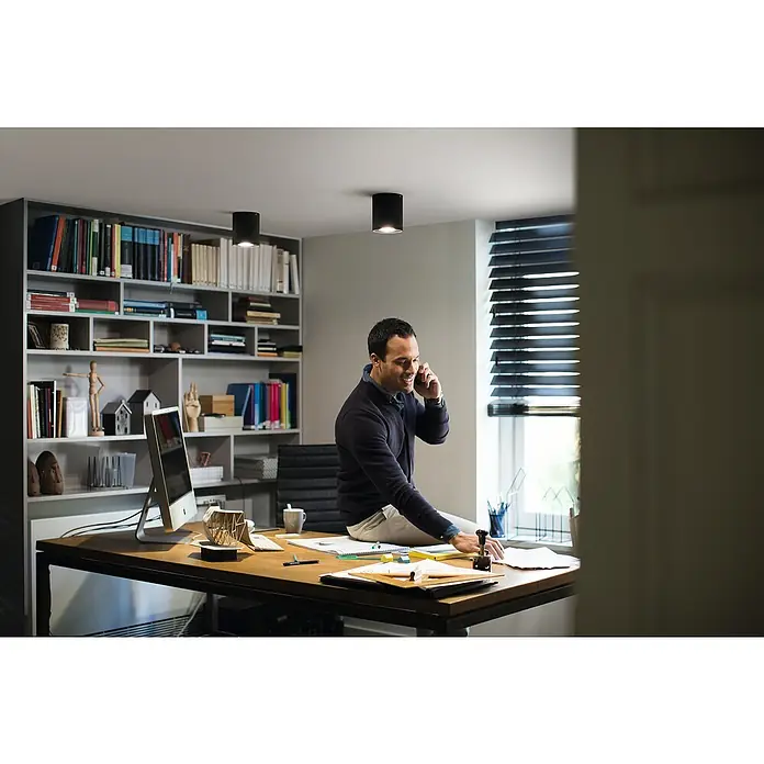 Philips Hue LED-Deckenstrahler White Ambiance Pillar - 4,2 W, L x B x H: 10,3 x 10,3 x 12 cm, Schwarz, Neutralweiß - Mann sitzt auf einem Tisch und telefoniert in einem Büro mit Beleuchtung.