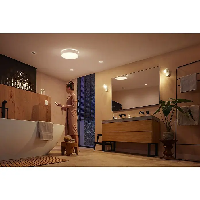 Philips Hue LED-Deckenleuchte rund Devere - 34 W, Ø x H: 47,6 x 10 cm, Weiß, Neutralweiß - Modernes Badezimmer mit einem smarten Deckenlicht und einer Frau im Bademantel, die ein Smartphone hält, um die Beleuchtung zu steuern.