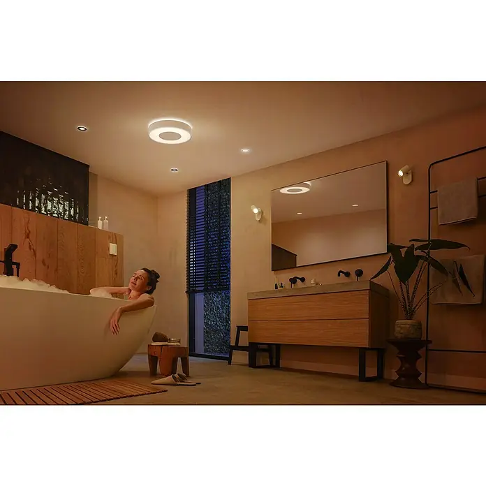 Philips Hue LED-Deckenleuchte rund Xamento - 53 W, Ø x H: 48,1 x 8,4 cm, Weiß, RGBW - Ein helles Badezimmer mit einer Frau in der Badewanne und einer smarten Philips Deckenleuchte, die eine entspannende Atmosphäre schafft.