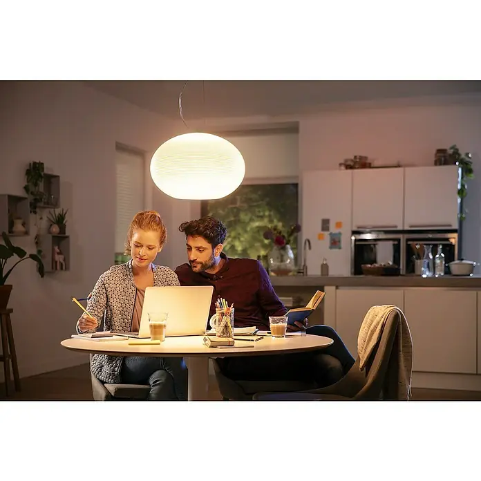 Philips Hue LED-Pendelleuchte Flourish - 39,5 W, L x B x H: 40,1 x 40,1 x 30 cm, Weiß - Ein Paar sitzt am Tisch unter einer runden Pendelleuchte. Die Leuchte ist hell und spendet warmes Licht, ideal für gemütliche Abende.