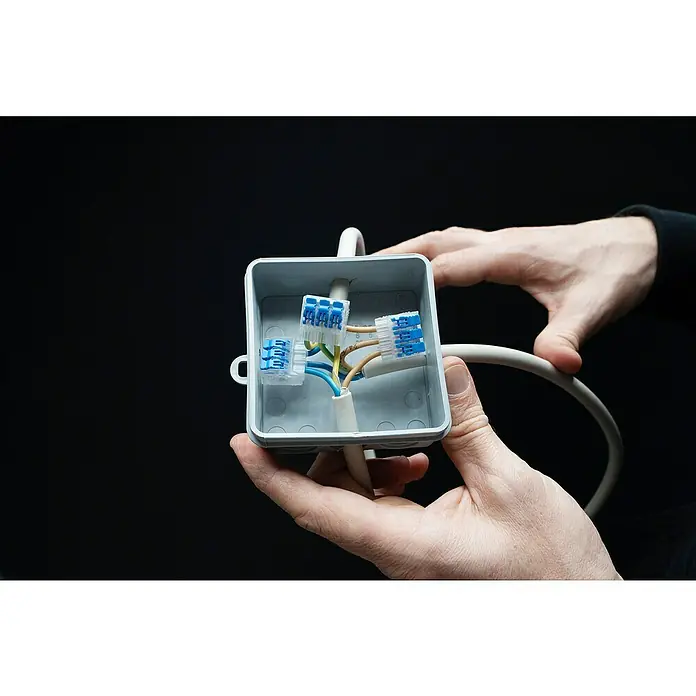 Ideal Hebelklemme Gen II - Anzahl Leiter: 3 Stk., 4 mm², 40 Stk. - Nahaufnahme einer grauweißen Verteilerdose mit blauen Wago-Klemmen, gehalten von zwei Händen vor einem schwarzen Hintergrund.