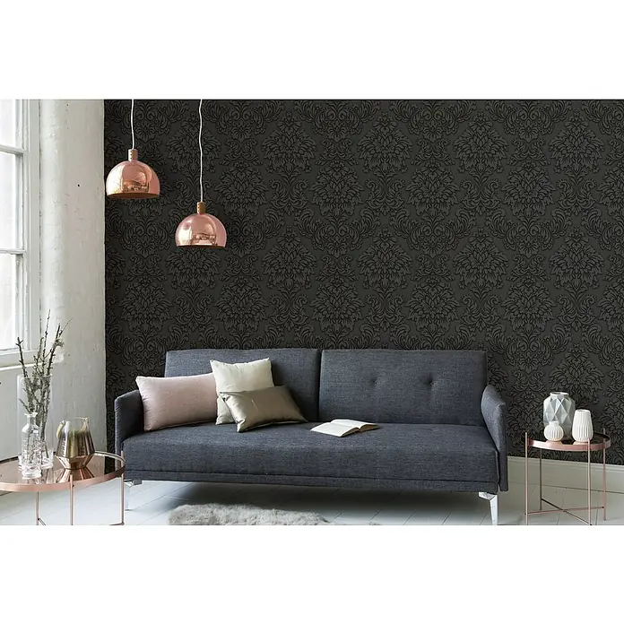 A.S. Création Metropolitan Stories Vliestapete Damask - Grau/Schwarz, Ornament, 10,05 x 0,53 m - Elegantes Interieur mit einem stilvollen grauen Sofa, akzentuiert durch Kupferleuchten und dekorative Kissen vor einer dunklen, gemusterten Tapete.