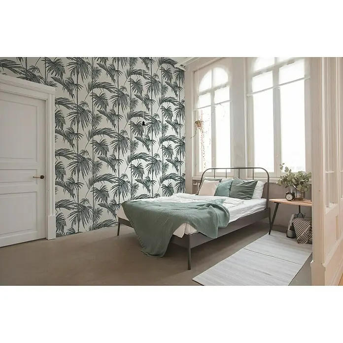 A.S. Création Metropolitan Stories Vliestapete Gräser - Weiß/Silber, Floral, 10,05 x 0,53 m - Einladendes Schlafzimmer mit einem Bett mit dunkelgrauem Rahmen und einer Wand mit Palmenmuster.
