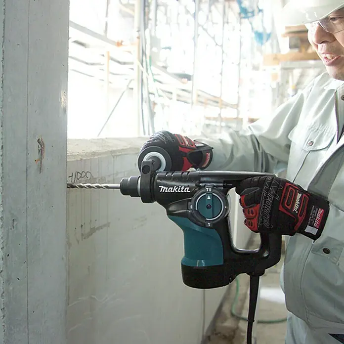 Makita Martillo perforador HR2810 - 800 W, Portabrocas de sujeción rápida - Un trabajador de la construcción perfora un agujero en una pared de hormigón con un martillo rotatorio Makita. La herramienta es negra y verde azulado.