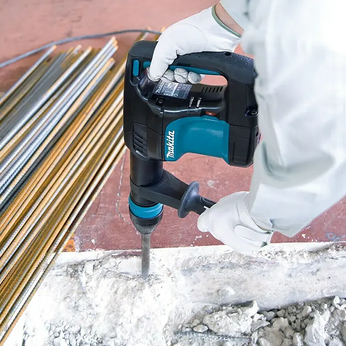 Makita Martillo percutor HM0870C - 1.100 W, Alojamiento SDS-MAX - Un martillo demoledor Makita en acción, operado por una persona con guantes blancos, rompiendo concreto. Herramienta y protección en armonía.