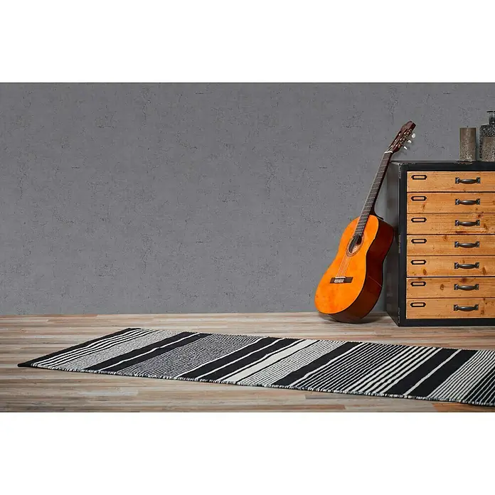 A.S. Création Metropolitan Stories Vliestapete Betonoptik - Dunkelgrau, Betonoptik, 10,05 x 0,53 m - Ein gestreifter Teppich in Schwarz und Weiß liegt auf einem Holzboden, neben einer Gitarre und einer Kommode. Die Streifen sind diagonal angeordnet.