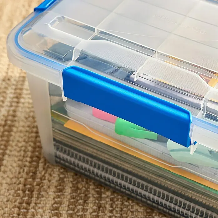 Aufbewahrungsbox Wasserdicht - L x B x H: 38,2 x 28,8 x 24,3 cm, Kunststoff, Transparent - Ein durchsichtiger Aufbewahrungsbehälter aus Kunststoff mit blauem Deckelverschluss, gefüllt mit Büromaterial und Heften, steht auf einem Teppich.