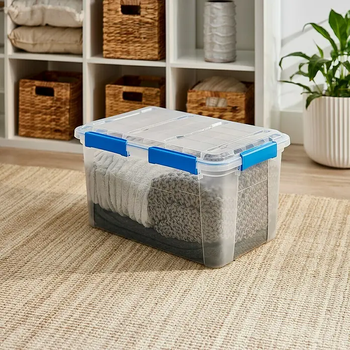 Aufbewahrungsbox Wasserdicht - L x B x H: 57,8 x 38,2 x 31,8 cm, Kunststoff, Transparent - Bild einer transparenten Aufbewahrungsbox mit blauem Verschluss, gefüllt mit Textilien, auf einem Teppich vor einem weißen Regal.