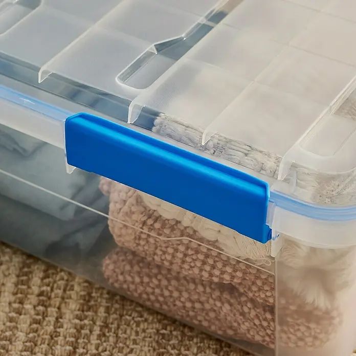 Aufbewahrungsbox Wasserdicht - L x B x H: 57,8 x 38,2 x 21,7 cm, Kunststoff, Transparent - Ein durchsichtiger Kunststoffbehälter mit blauem Verschlussclip, gefüllt mit Textilien, steht auf einem Teppich.