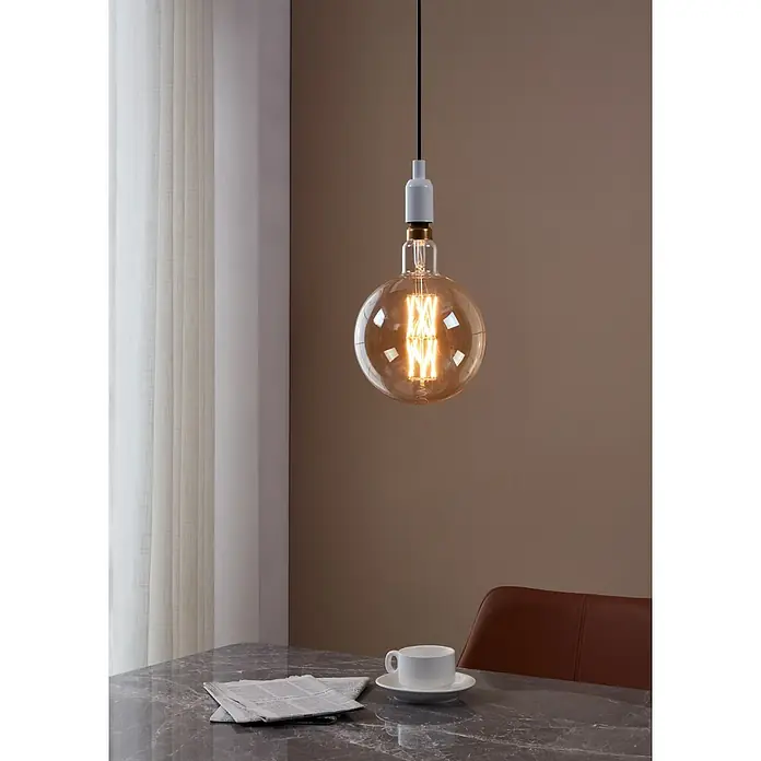 Eglo LED-Lampe - E27, Dimmbar, Warmweiß, 806 lm, 8 W - Nahaufnahme einer Edison-Glühbirne, die von der Decke hängt und ein warmes Licht über einem Tisch mit einer Tasse und einer Zeitung spendet.