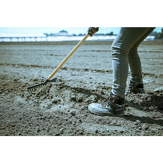 Bellota Rastrillo de jardín - Anchura de trabajo: 36 cm, Número de púas: 16 - Primer plano de una persona rastrillando la tierra, destacando las robustas botas de trabajo Bellota diseñadas para condiciones difíciles.
