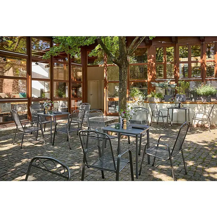 Sunfun Gastro Gartentisch - L x B x H: 74,5 x 75 x 74,5 cm, Stahl, Night-Grey, Stapelbar - Außenaufnahme von grauen Metalltischen und Stühlen auf einer Terrasse mit Baumbeschattung, ideal für entspannte Stunden im Freien.