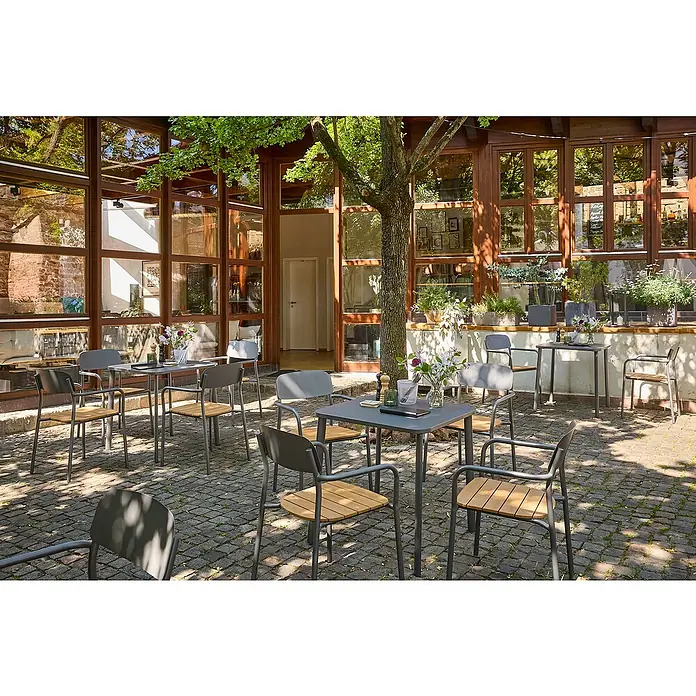Sunfun Gastro Gartentisch - L x B x H: 74,5 x 75 x 74,5 cm, Stahl, Night-Grey, Stapelbar - Stühle und Tische im Freien vor einem Gebäude mit großen Fenstern. Die grauen Stühle haben hölzerne Sitze und verleihen der Szene einen Hauch von Eleganz.