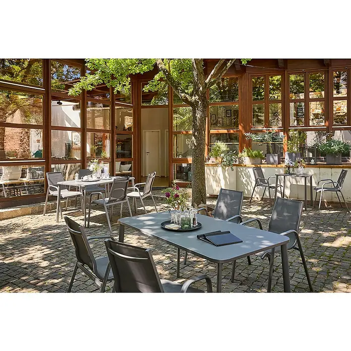 Sunfun Gastro Gartentisch - L x B x H: 142,5 x 82,5 x 75 cm, Stahl, Night-Grey, Stapelbar - Ein einladender Außenbereich mit grauen Tischen und Stühlen. Perfekt für Mahlzeiten im Freien, umgeben von üppigem Grün und natürlichem Licht.