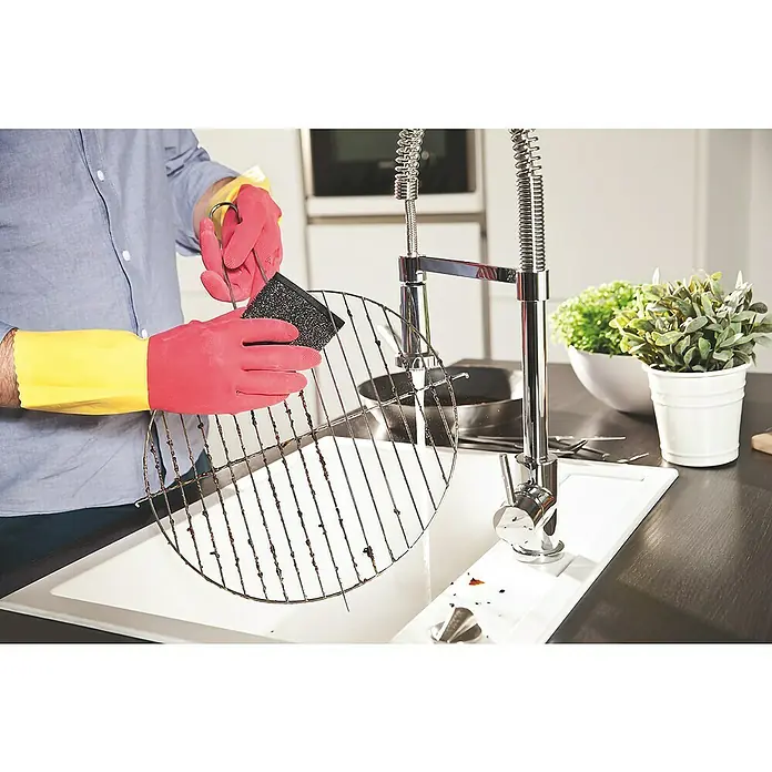 Vileda Glitzi Reinigungspad Power Inox - 2 Stk. - Bild einer Person, die ein schmutziges Grillrost mit einem Scheuerschwamm und Gummihandschuhen in einer Küchenspüle reinigt.