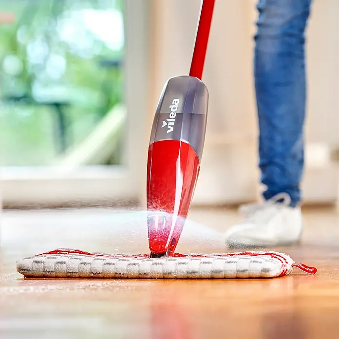 Vileda Bodenwischer Spray & Clean Sprühwischer - 2 -tlg. - Nahaufnahme eines Vileda-Sprühmopps in Aktion auf einem Holzboden, der Reinigungsflüssigkeit versprüht, mit unscharfem Hintergrund.
