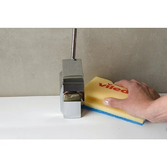 Vileda Glitzi Schwamm Jumbo sanft - Glitzi Härtegrad: 1, 1 Stk. - Eine Hand reinigt einen modernen Wasserhahn mit einem gelb-blauen Vileda-Schwamm auf einem weißen Waschbecken, vor einem grauen Hintergrund.