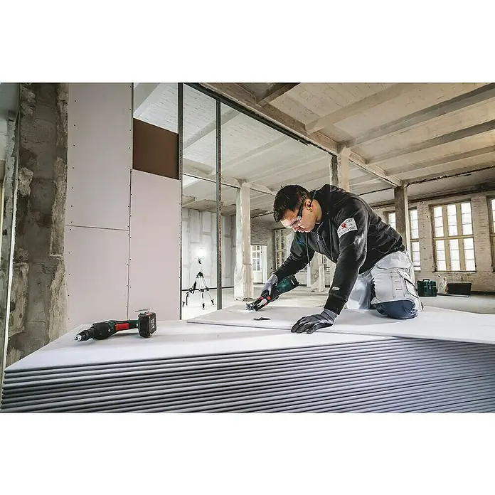 Metabo CAS 18V Akku-Multifunktionswerkzeug MT 18 LTX - 18 V - Ein Handwerker, der mit einem Elektrowerkzeug eine Gipskartonplatte bearbeitet, während ein Akkuschrauber daneben liegt. Im Hintergrund ist ein Rohbau zu sehen.