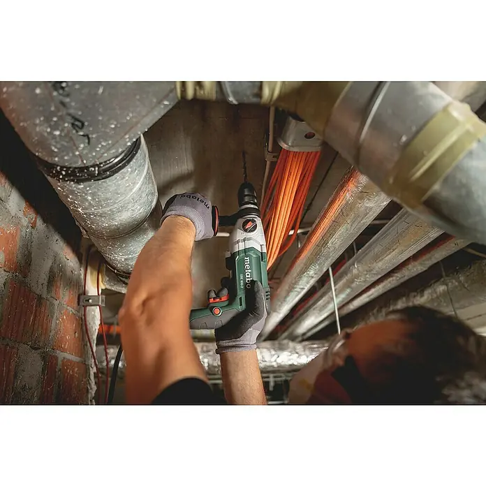 Metabo Schlagbohrmaschine - 800 W, Schnellspannbohrfutter, Max. Schlagzahl: 51.200 /min - Ein Mann bohrt mit einer Metabo SBE 800-2 Schlagbohrmaschine ein Loch in eine Betondecke, umgeben von Rohren und Kabeln. Er trägt Handschuhe und eine Schutzbrille.