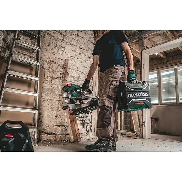 Metabo CAS 18V Akku-Baustellenradio RC 12-18 32 W BT DAB+ - 18 V, Ohne Akku, 87,5 - 108,0 kHz (FM) - Ein Mann trägt eine Metabo Kapp- und Gehrungssäge und einen Metabo Lautsprecher in einem unrenovierten Raum.