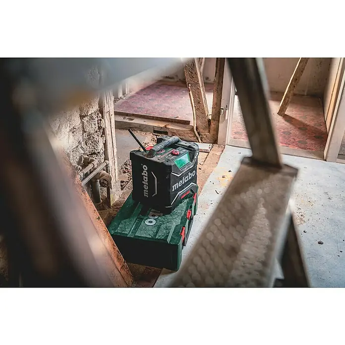 Metabo CAS 18V Radio de batería para la obra RC 12-18 DAB+ - 18 V, Sin batería, 87,5 - 108,0 kHz (FM) - Radio de obra Metabo sobre una caja de herramientas en una habitación que necesita renovación. La radio es negra con detalles verdes y tiene una antena.