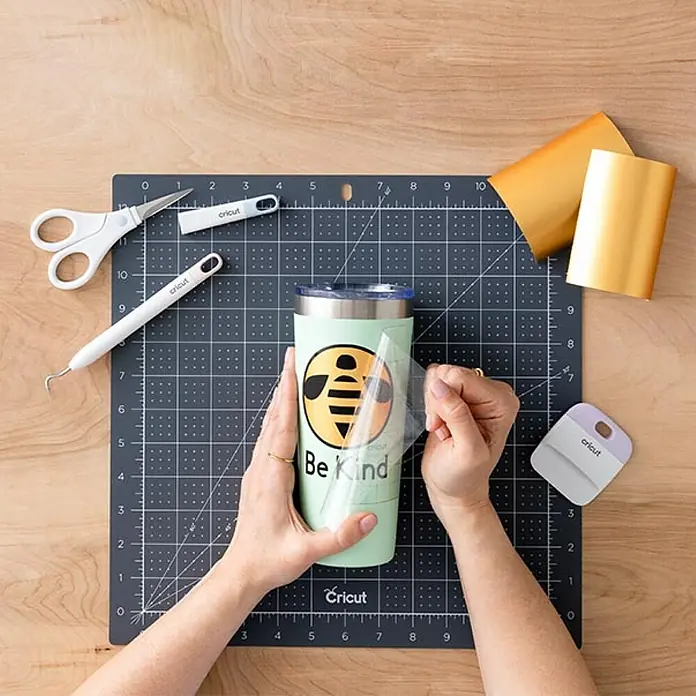 Cricut Transferfolie Transfer Tape - L x B: 121,9 x 30,5 cm, Passend für: Cricut Maker - Nahaufnahme eines Cricut-Tassenprojekts mit der Aufschrift "Be Kind", Werkzeugen und Materialien auf einer Schneidematte.