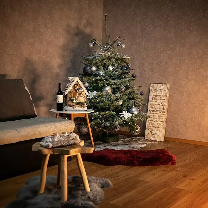 Piardino Nordmanntanne - 150 - 200 cm hoch, Abies Nordmanniana - Ein geschmückter Weihnachtsbaum mit silbernen und blauen Kugeln und Lichtern, der eine gemütliche Winterecke mit Möbeln und festlicher Dekoration schmückt.