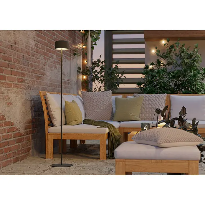 Eglo Solar-Stehleuchte Roccanova-L - Grün, IP44, Ø x H: 13,5 x 120 cm - Eine moderne Stehlampe aus schwarzem Metall steht neben einem Lounge-Set mit Holztisch und gepolsterten Kissen im Außenbereich.