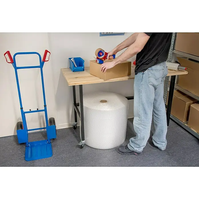 Rikta Werkbank mit Rollen - L x B: 140 x 60 cm, Arbeitshöhe: 84 cm, Belastbarkeit: 200 kg - Ein blauer Rollwagen steht neben einem Tisch, auf dem eine Person eine braune Kiste mit einem Handabroller verpackt. Eine Rolle Luftpolsterfolie darunter.