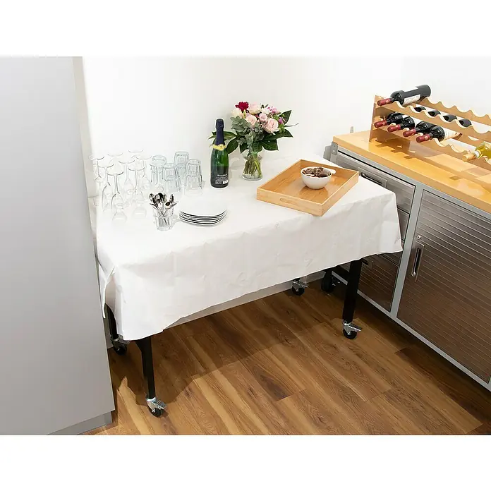 Rikta Werkbank mit Rollen - L x B: 140 x 60 cm, Arbeitshöhe: 84 cm, Belastbarkeit: 200 kg - Ein Tisch mit Rädern, gedeckt mit einer weißen Tischdecke, auf der Gläser, Teller und Getränke stehen.