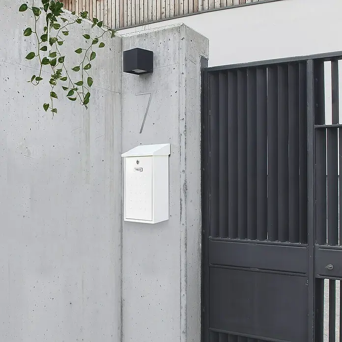 Arregui Premium Buzón de pared E-5601 - L x An x Al: 25 x 10 x 40 cm, Blanco, Acero - La imagen muestra un buzón blanco moderno montado en una pared de hormigón gris, junto a una puerta metálica oscura.