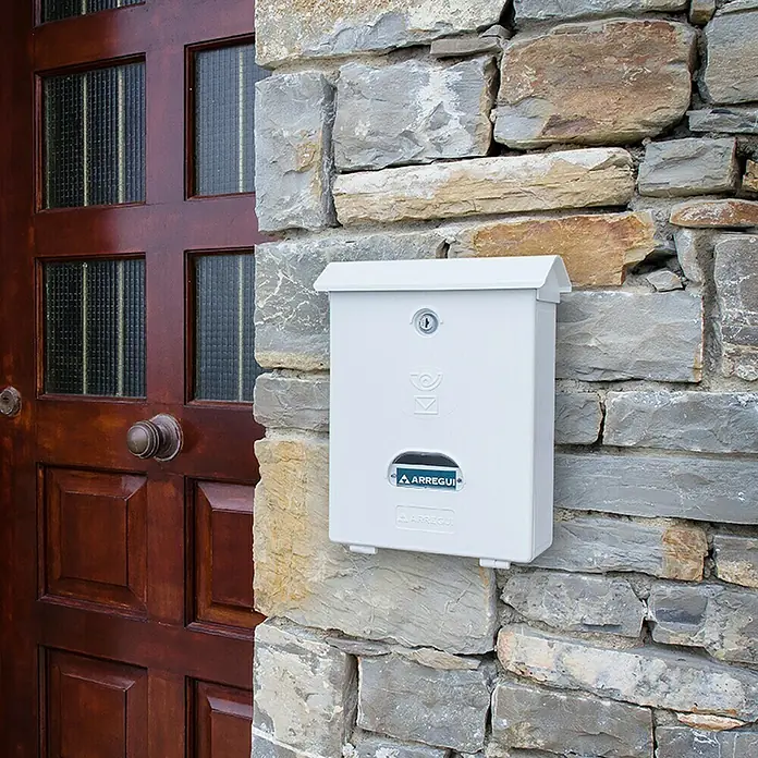 Arregui Classic Buzón de pared E-107 - L x An x Al: 27 x 11 x 34,6 cm, Blanco, Plástico - Primer plano de un buzón blanco Arregui montado en una pared de piedra junto a una puerta de madera. Diseño sencillo y funcional.