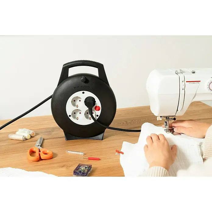 Brennenstuhl Kabeltrommel Vario Line VL2-X - Kunststoff, 10 m, H05VV-F, IP20 - Schwarze Kabeltrommel mit vier Steckdosen und eingestecktem Stecker auf einem Tisch neben einer Nähmaschine.