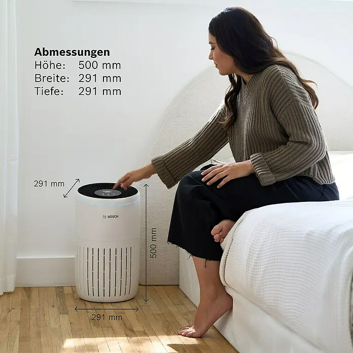 Bosch Luftreiniger Air 4000 - Raumgröße: 62,5 m², 29,1 x 50 cm - Eine Frau sitzt auf einem Bett und bedient einen Bosch Luftreiniger. Die Abmessungen des Geräts sind angegeben.