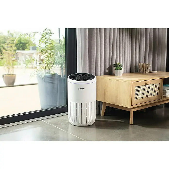 Bosch Luftreiniger Air 4000 - Raumgröße: 62,5 m², 29,1 x 50 cm - Ein Bosch Luftreiniger steht in einem modernen Wohnzimmer, neben einem Fenster und einem Holzschrank, für saubere Luft und ein gesundes Raumklima.