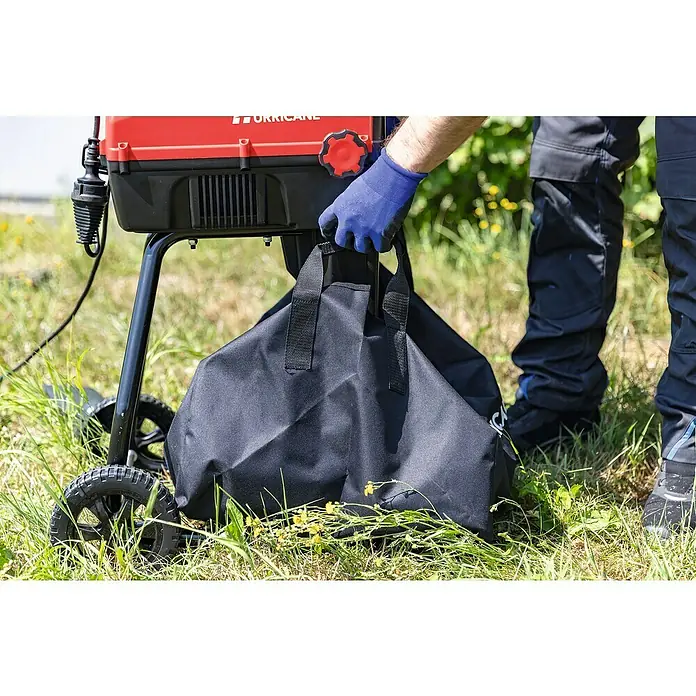 Hurricane Elektro-Leisehäcksler HLH-S-2800 - Max. Aststärke: 44 mm, 2.800 W - Eine Nahaufnahme eines Häckslers mit einem blauen Arbeitshandschuh, der eine schwarze Tasche hält, mit einer Person in Arbeitskleidung im Hintergrund.