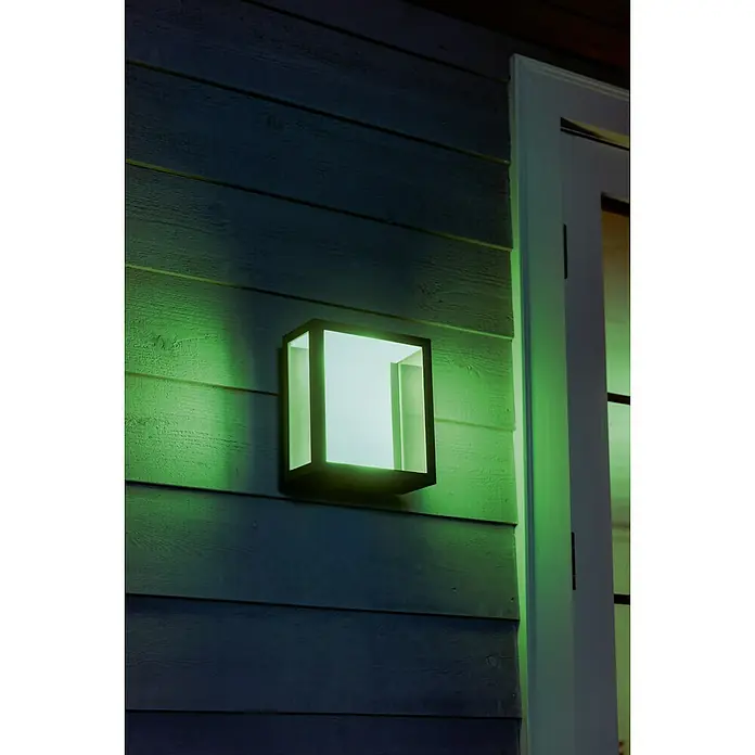 Philips Hue LED-Außenwandleuchte Impress - 8 W, 11,7 x 19 x 24 cm, Schwarz, IP44 - Eine quadratische Außenleuchte mit schwarzem Rahmen und grünem Licht ist an einer Holzfassade neben einer Tür montiert.