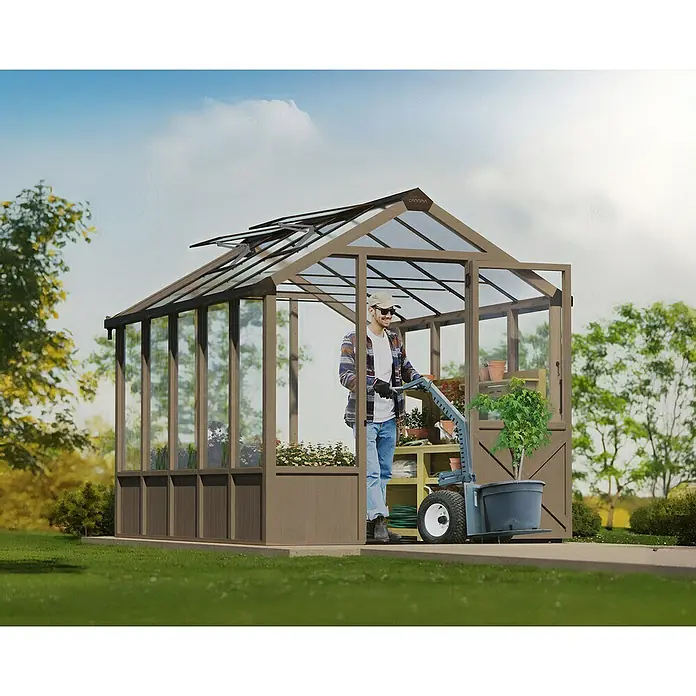 Palram – Canopia Invernadero Natura - Medidas ext. (An x Pr): 249 x 356 cm, Plástico, 4 mm, Natural - Hombre con carro transportador de plantas frente a un invernadero. El invernadero ofrece espacio para plantas y verduras y alarga la temporada.