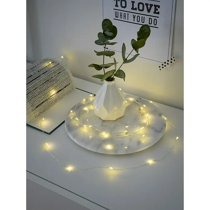 Konstsmide LED-Lichterkette - Innen, Lichtfarbe: Warmweiß, Anzahl Leuchtmittel: 20 Stk., Batteriebetrieben, Silber - Dekorative LED-Lichterkette, arrangiert um eine Vase und ein Buch auf einem weißen Tisch, für eine gemütliche Atmosphäre.